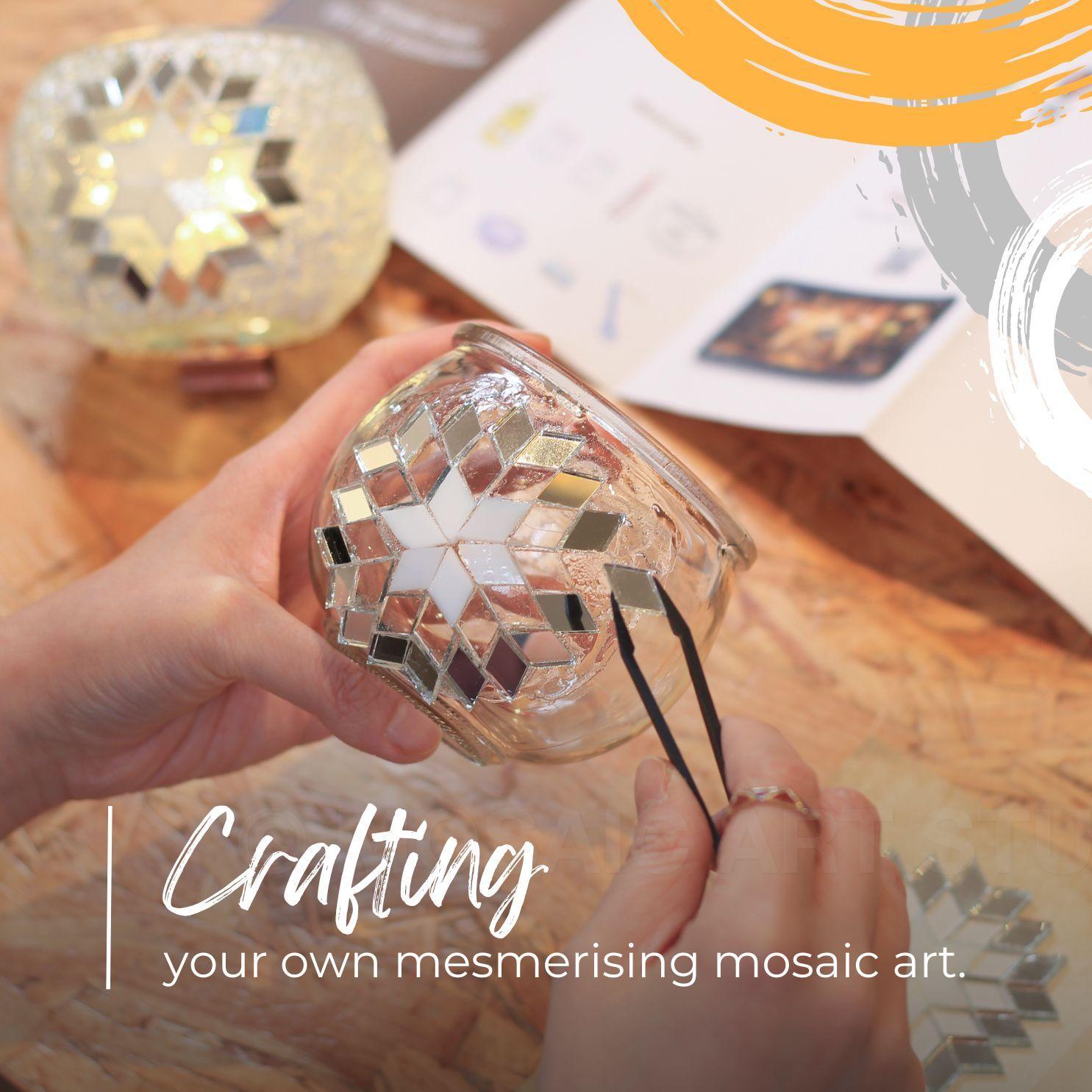 Hands crafting a mosaic candle holder with mirrored and white glass tiles using tweezers on wooden surface