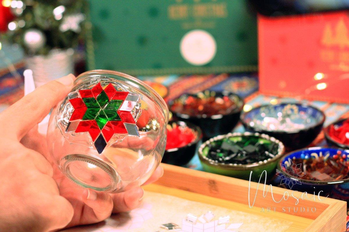 Hand holding glass candle holder decorated with red and green mosaic star in art studio DIY kit