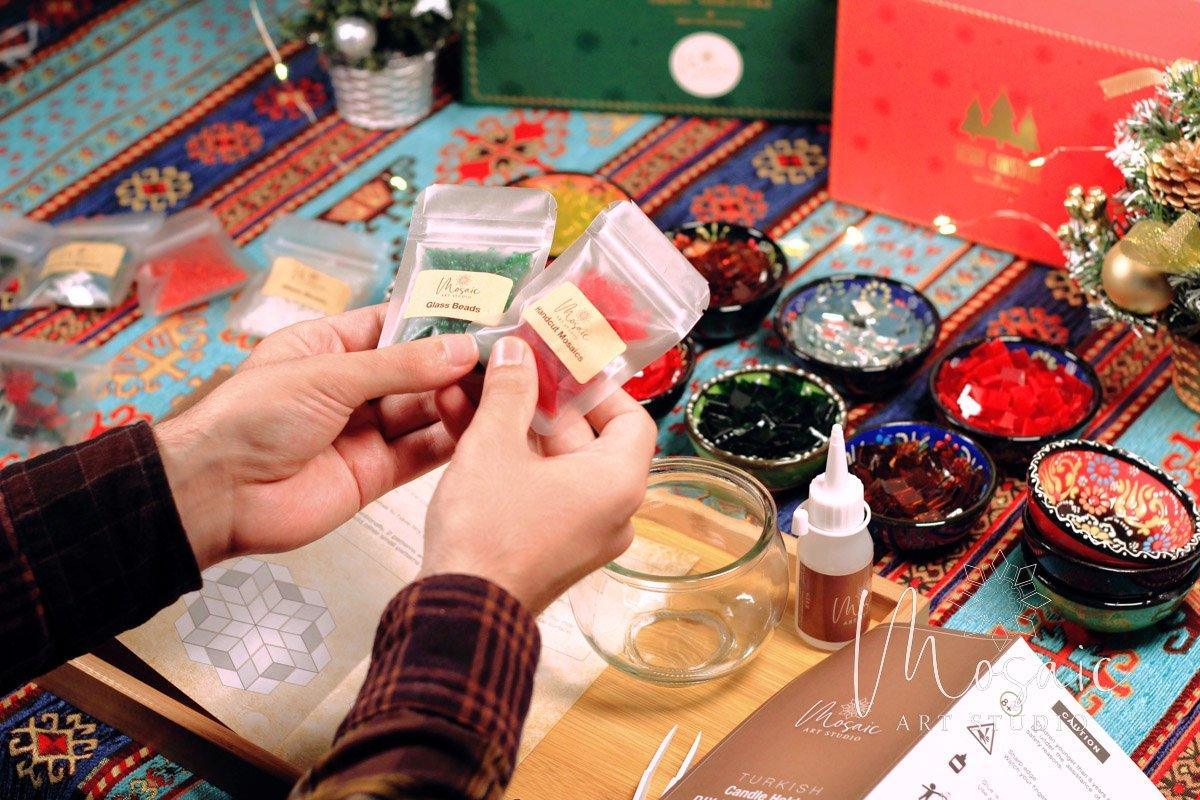 Hands holding packets of green glass beads and red handcut mosaics for DIY mosaic candle holder kit