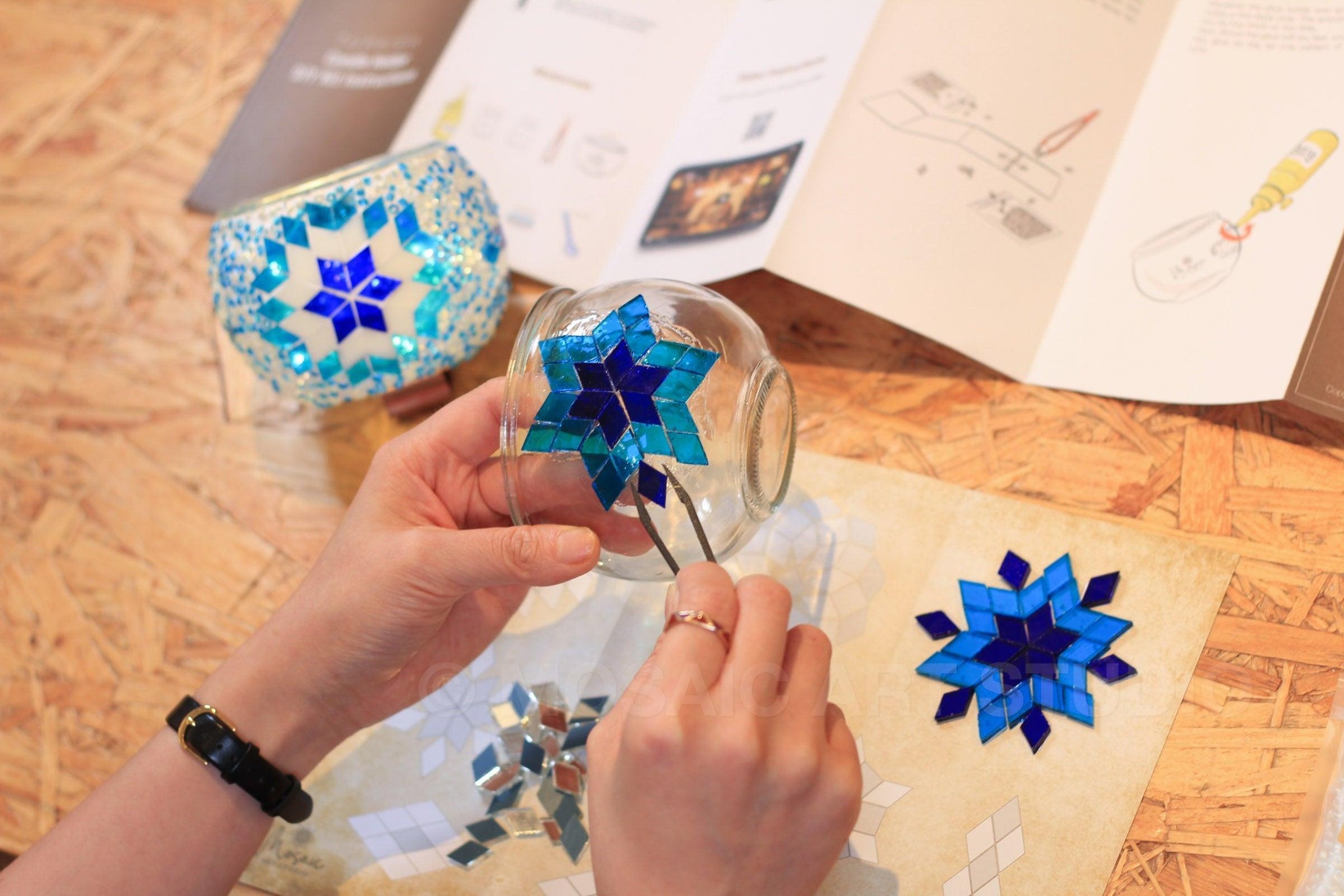 Hands using tweezers to apply blue mosaic pieces on glass candle holder, DIY mosaic craft kit