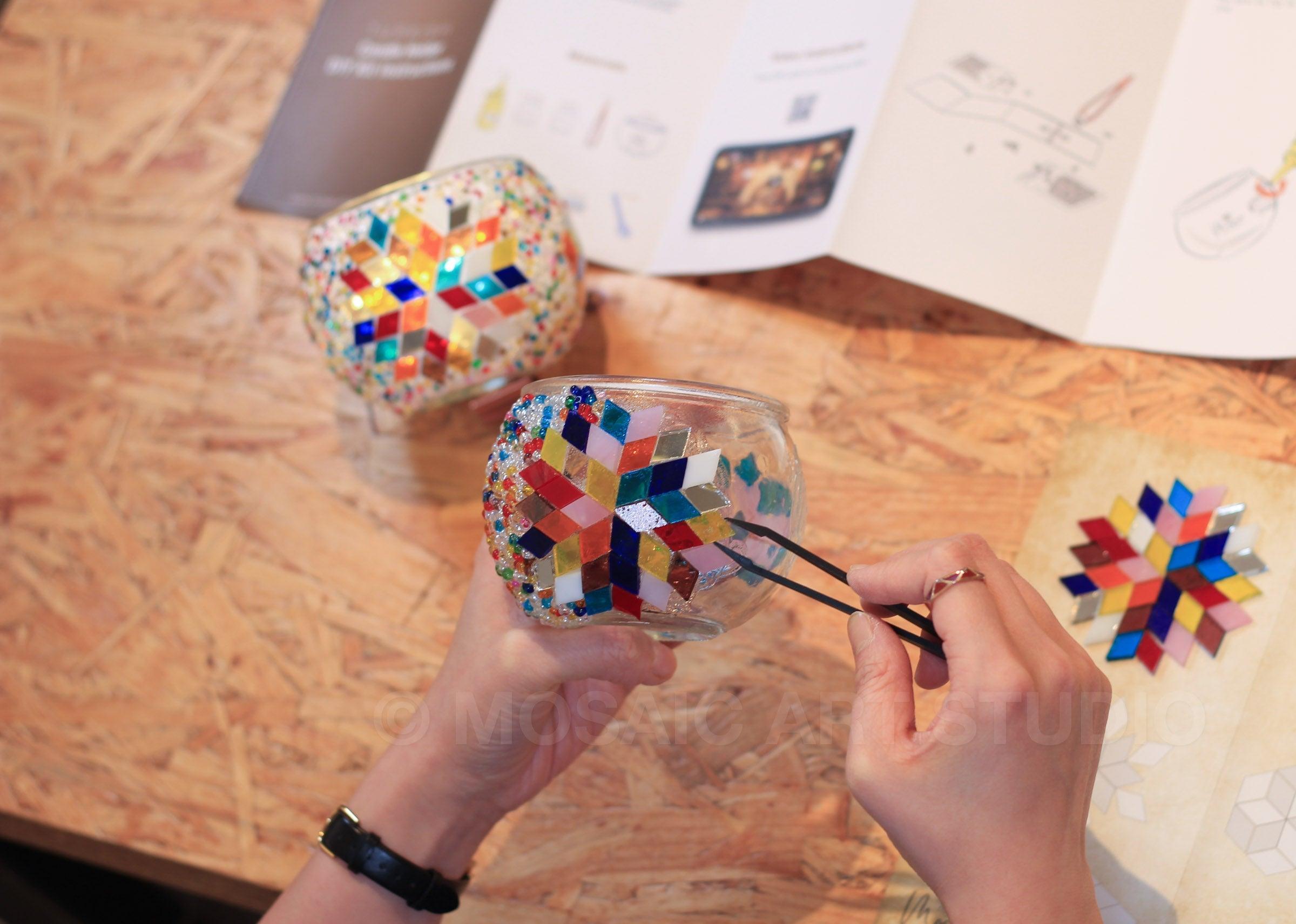 Hands decorating glass candle holder with colorful mosaic tiles using tweezers on wooden table