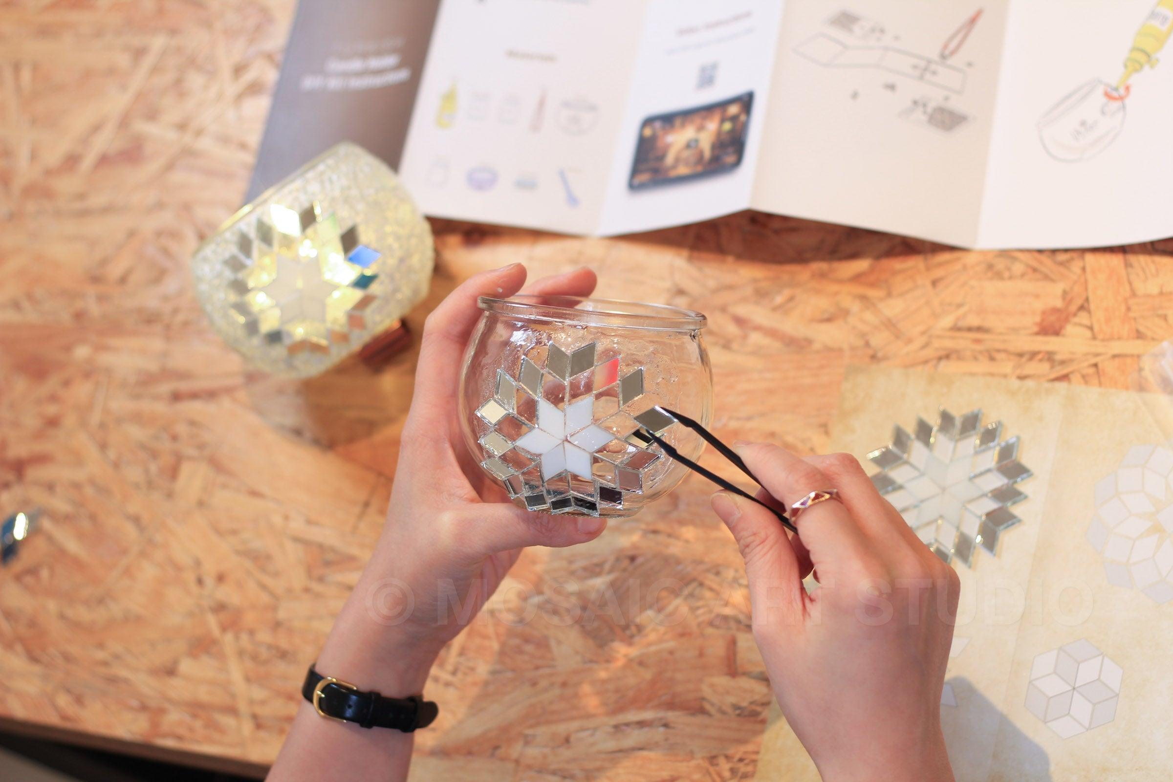 Hands assembling mosaic mirror pieces on glass candle holder DIY kit with instruction sheet on wooden table