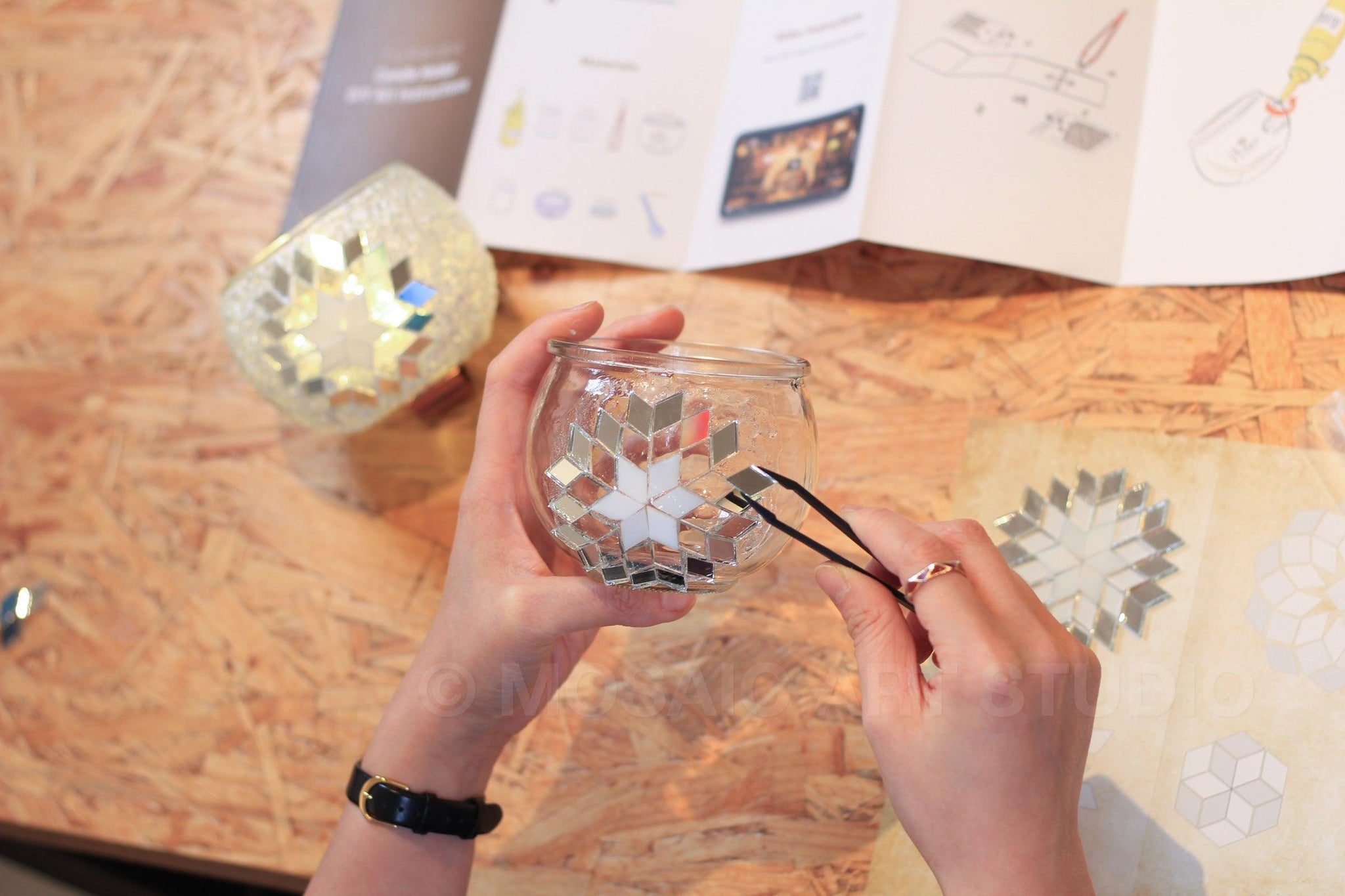 Hands assembling mosaic mirror pieces on glass candle holder DIY kit with instruction sheet on wooden table