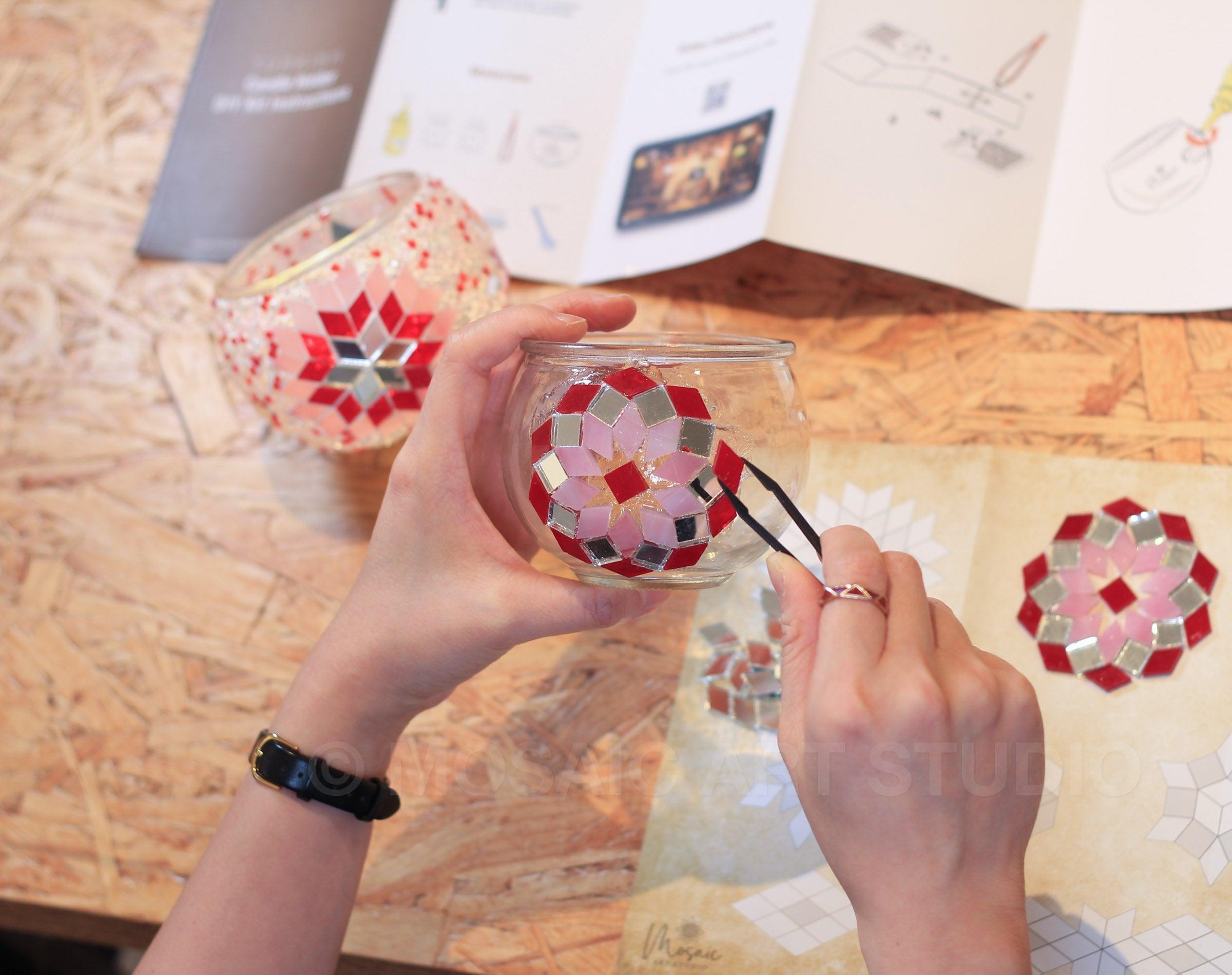 Hands creating a mosaic candle holder with red, pink, and mirror tiles using tweezers on a wooden table